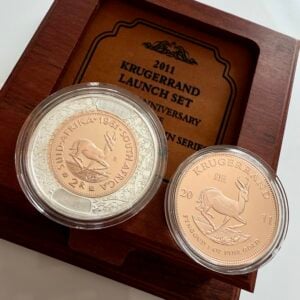 Krugerrand 2011 1 oz Gold Proof in transparenter Kapsel; Rückseite mit Springbock, Umschrift „KRUGERRAND – FYNGOUD 1 OZ FINE GOLD – 2011“, im Hintergrund Holzetui mit „Krugerrand Launch Set – 50 Year Anniversary of the First Decimal Coin Series“.