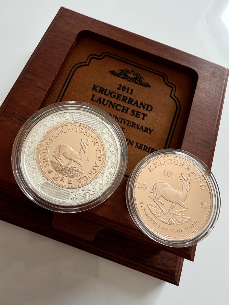 Krugerrand 2011 1 oz Gold Proof in a transparent capsule; reverse with a springbok, inscription “KRUGERRAND – FYNGOUD 1 OZ FINE GOLD – 2011”, in the background a wooden case with “Krugerrand Launch Set – 50 Year Anniversary of the First Decimal Coin Series”.