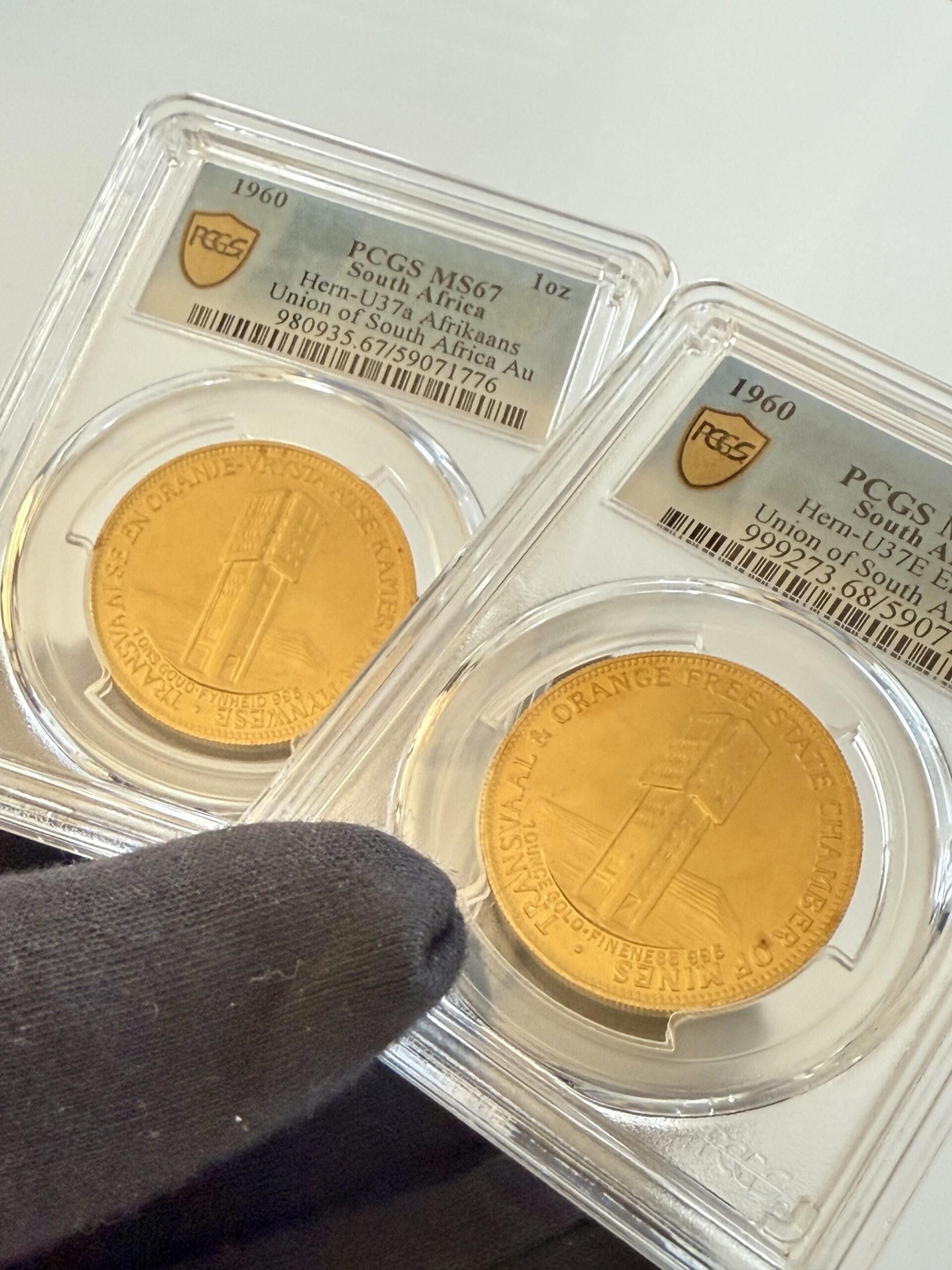suedafrika-1960-chamber-of-mines-1oz-gold-afrikaans-und-englisch.pcgs_1 Zwei PCGS-Slabs nebeneinander mit Südafrika 1960 Chamber of Mines 1oz Gold. Links Afrikaans-Variante Hern-U37a PCGS MS67, rechts English-Variante Hern-U37b PCGS MS68, jeweils mit Turmmotiv.