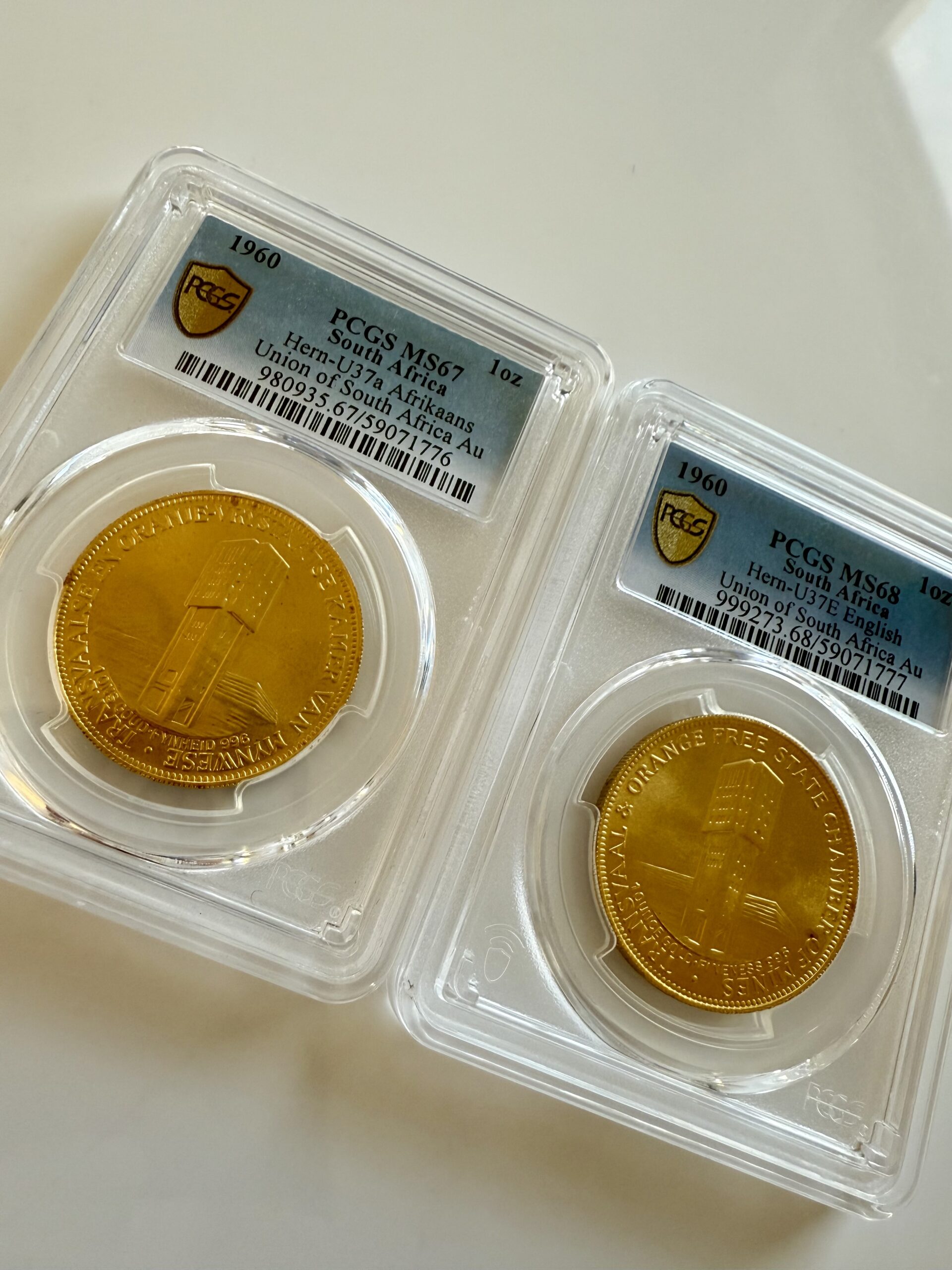 suedafrika-1960-chamber-of-mines-1oz-gold-afrikaans-und-englisch.pcgs_4 Zwei PCGS-Slabs nebeneinander mit Südafrika 1960 Chamber of Mines 1oz Gold. Links Afrikaans-Variante Hern-U37a PCGS MS67, rechts English-Variante Hern-U37b PCGS MS68, jeweils mit Turmmotiv.