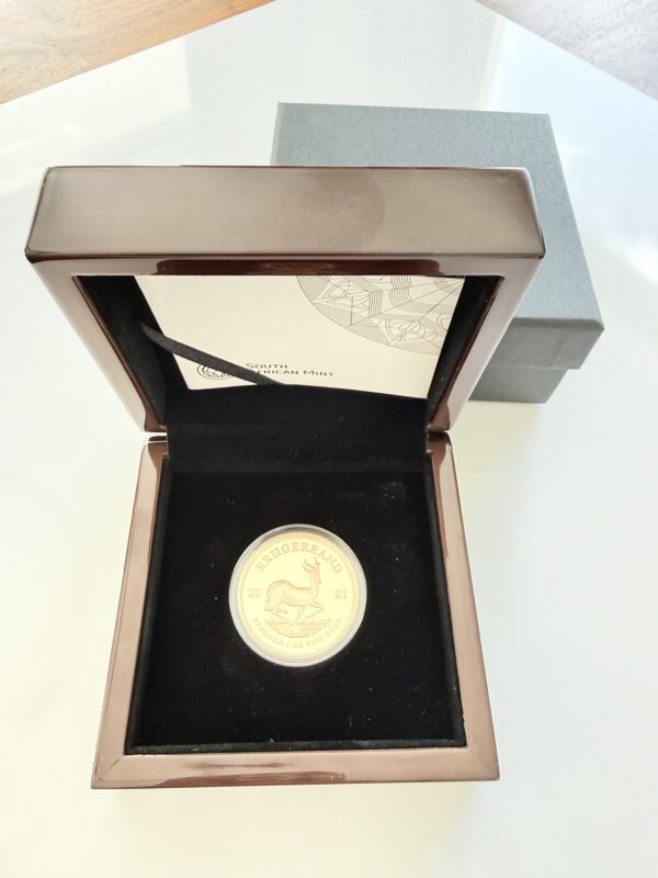 Südafrikanische Krügerrand Goldmünze von 2021 in Kapsel, fotografiert vor ihrer Originalverpackung. Zu sehen ist die Springbock-Antilope mit der Jahreszahl 2021 sowie der Umschrift „Krugerrand“ und „Fyngoud 1 oz Fine Gold“.