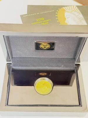 usa-2019-100-dollars-american-liberty-gold-high-relief-enhanced-uncirculated-1oz_2 Moneta d'oro American Liberty del 2019 in una confezione di presentazione aperta con custodia nera, capsula inserita e certificato di autenticità parzialmente visibile sullo sfondo.