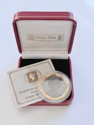 Isle of Man 1990 Crown Gold 150 Years Penny Black_2 Original presentation case of an Isle of Man gold coin with the red presentation box open, containing the coin in a capsule and an accompanying certificate. The coin is inscribed "Penny Black - 150th Anniversary - Crown", the certificate "Certificate of Proof Penny Black", and the lid reads "Pobjoy Mint - England UK".