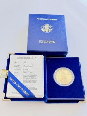 Estuche original de una moneda de oro American Eagle de 1987 con la caja de presentación azul abierta, que contiene una moneda de oro proof de 1 onza en una cápsula y una hoja de especificaciones adjunta de la Casa de la Moneda de los Estados Unidos. Al fondo se ve la caja exterior azul a juego con la inscripción "American Eagle - One Ounce Proof Gold Bullion Coin".