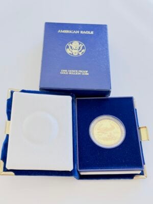 EE. UU. 1987 American Eagle Gold 1oz proof_2 Estuche original de una moneda de oro American Eagle de 1987 con la caja de presentación azul abierta, que contiene una moneda de oro proof de 1 onza en una cápsula y una hoja de especificaciones adjunta de la Casa de la Moneda de los Estados Unidos. Al fondo se ve la caja exterior azul a juego con la inscripción "American Eagle - One Ounce Proof Gold Bullion Coin".